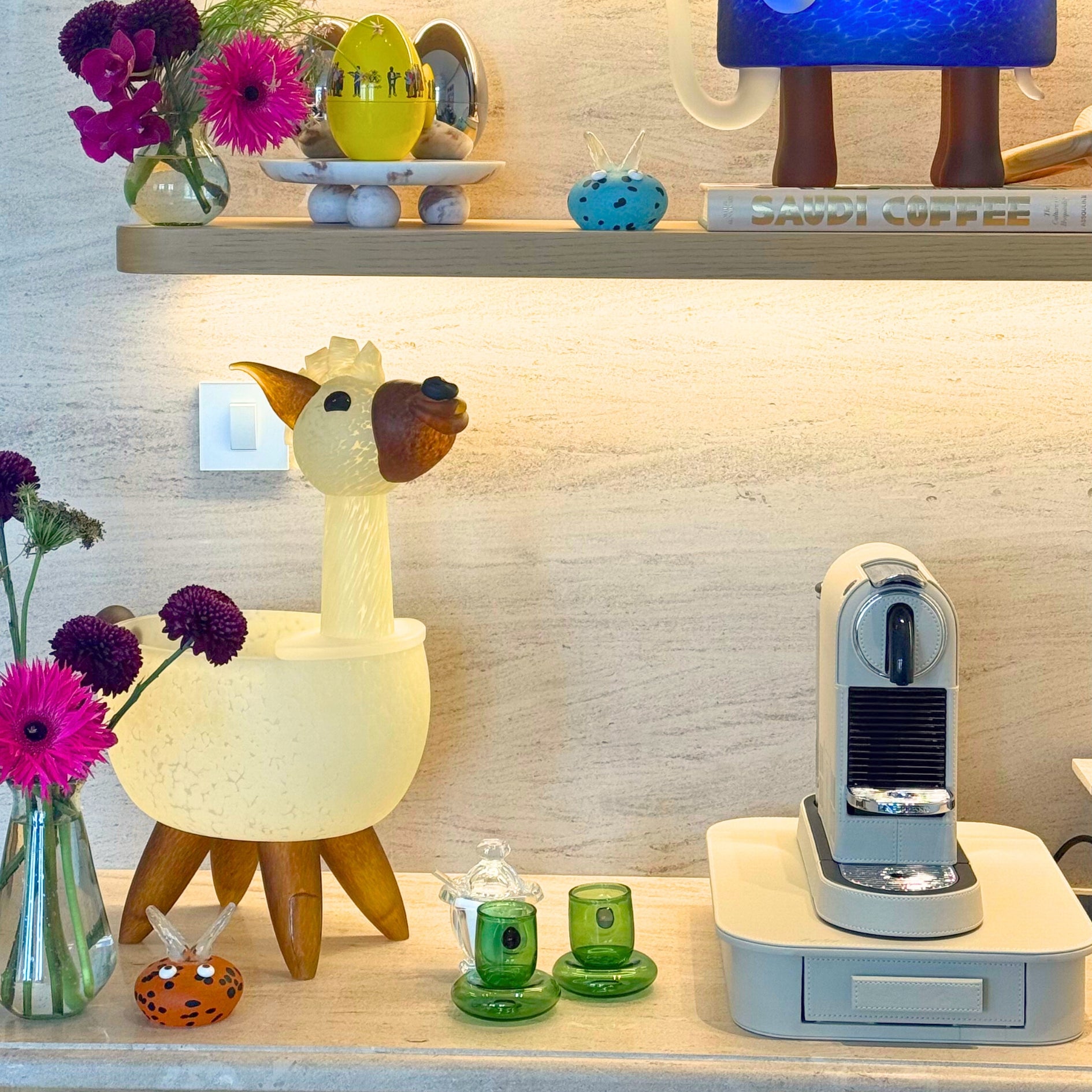 Decorative setup with a llama-shaped lamp, coffee machine, and flowers on a shelf.